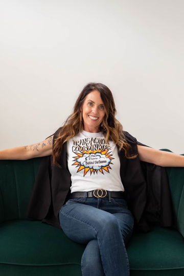 Jules seated on an emerald velvet couch in a black cape blazer over a Transaction Coordinator tee, arms open along the couch back, smiling at the camera.