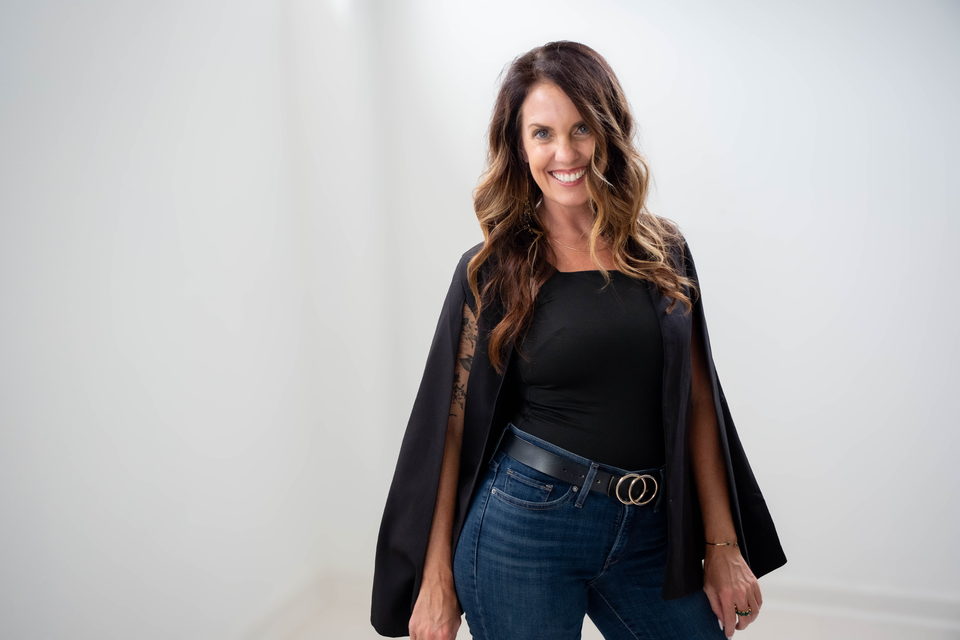 Julie Bowman in a black cape blazer, standing against a white studio wall, smiling directly at the camera.